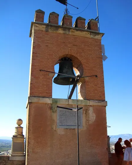 La campana de la Torre de la Vela de la Alhambra, colgada bajo su arco de ladrillo, recortada contra el cielo azul.