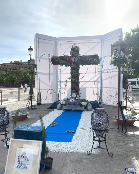 Cruz cubierta de claveles montada en una plaza, con cerámica azul y blanca, macetas y una alfombra azul a sus pies.