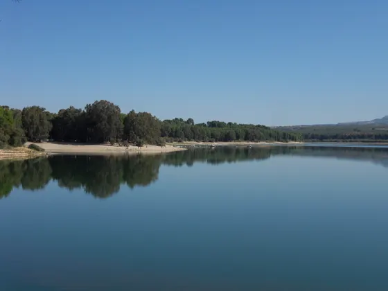 Orilla de arena del embalse de Cubillas con árboles hasta el agua, gente en la playa y el agua en calma, con el cielo reflejado.