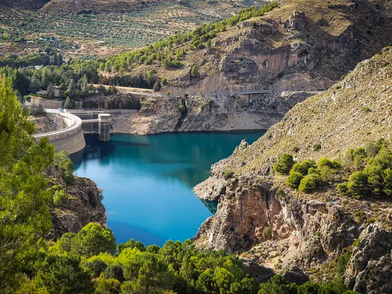 El embalse de Quéntar visto desde arriba, de agua turquesa entre laderas de roca y pinos, con la presa y su torre a la izquierda.