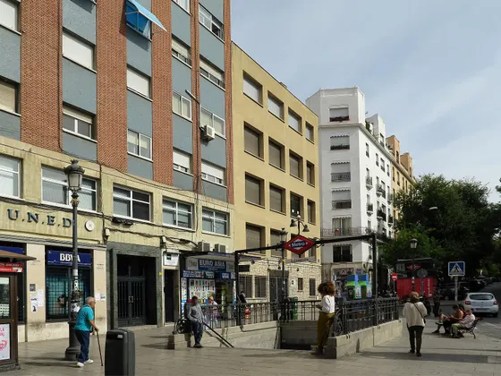 La plaza de Lavapiés con la entrada del metro en el centro, la sede de la UNED a la izquierda y gente cruzando la plaza.
