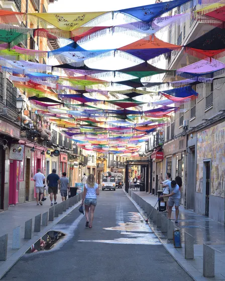 La Cava Baja vista a lo largo, con toldos de papel de colores cruzados entre los balcones y gente andando por el medio de la calle.