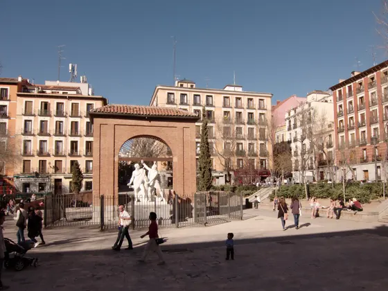 La plaza del Dos de Mayo con el arco de ladrillo del antiguo cuartel de Monteleón y el monumento a Daoíz y Velarde, y gente cruzando la plaza.